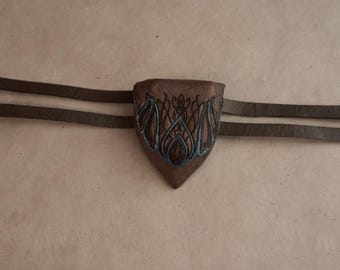 May include: A brown leather pouch with a triangular shape and a pointed bottom, featuring a dark blue intricate line-art design on the front. The pouch is attached to two thin brown leather straps that extend horizontally from both sides.