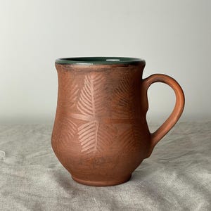 May include: A handmade, terracotta-colored ceramic mug with a leaf pattern. The mug has a dark green interior and a curved handle. The exterior features an embossed fern design. The mug is approximately 4 inches tall.