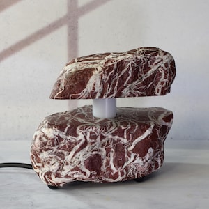 Puede incluir: Una base de lámpara de mármol rojo y blanco con un cable blanco. La base de la lámpara tiene forma de dos rocas apiladas.