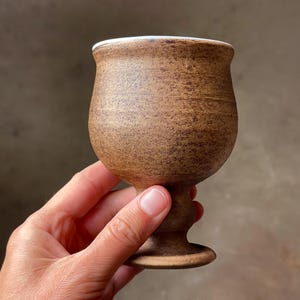 May include: A handmade, brown ceramic goblet with a textured surface and a white rim. The goblet has a rounded bowl, a stem, and a circular base. The overall design is rustic and earthy, suitable for beverages.