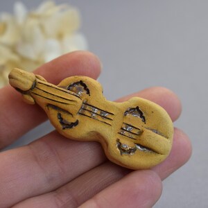 May include: A small, hand-painted ceramic violin figurine in a light brown color. The violin has a detailed design and is a charming decorative piece.