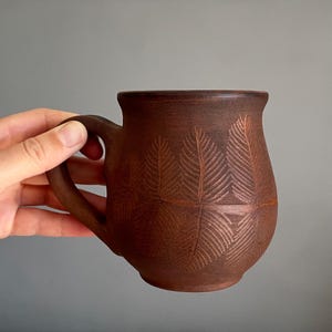 May include: A handmade brown ceramic mug with a leaf pattern. The mug has a rounded body, a sturdy handle, and a dark brown color. The leaf pattern is etched into the surface of the mug.