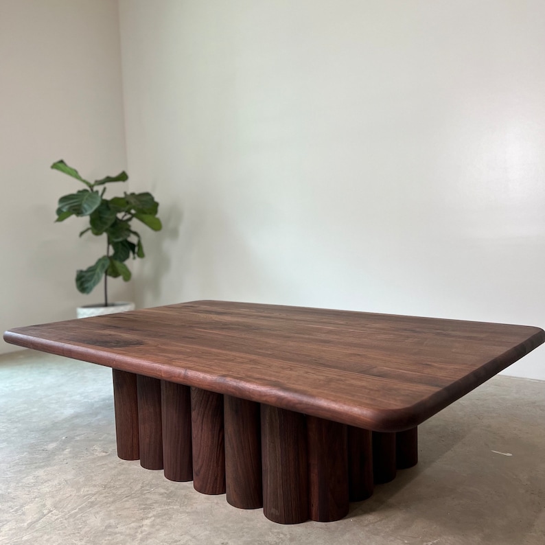 May include: A dark brown wooden coffee table with a rectangular top and a unique base made of stacked cylindrical pieces. The table is sitting on a light gray concrete floor.