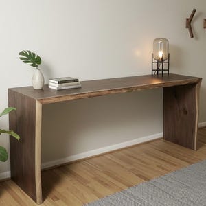 May include: A modern, dark wood console table with a live edge. The table has a natural wood grain and is paired with a black metal lamp, a vase with a leaf, and a potted plant. The table is approximately 72 inches long.