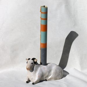 May include: A ceramic sheep candle holder with a striped candle. The sheep is white with black markings and is lying down. The candle is blue, orange, and grey. A unique decorative item.