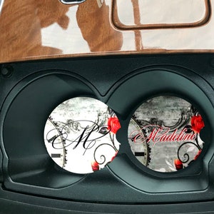 May include: Two round car coasters with a white background and a black and white floral design. One coaster has the letter "M" in black script and the other coaster has the name "Madeline" in black script. Both coasters have red roses.