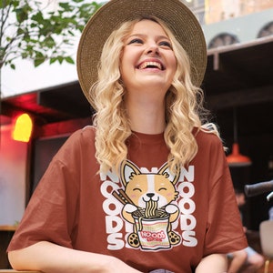 May include: A woman wearing a brown t-shirt with a cartoon corgi eating noodles. The shirt has the text "NOODS" repeated on it.