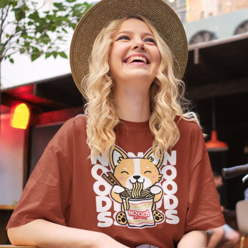 May include: A woman wearing a brown t-shirt with a cartoon corgi eating noodles. The shirt has the text "NOODS" repeated on it.