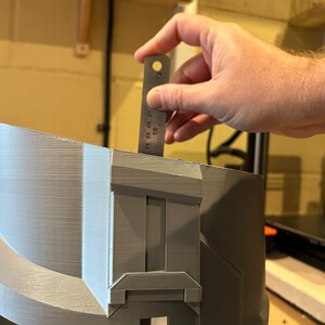May include: A gray 3D printed helmet with a detailed design. A ruler is being used to measure the helmet, which is approximately 7 inches tall.