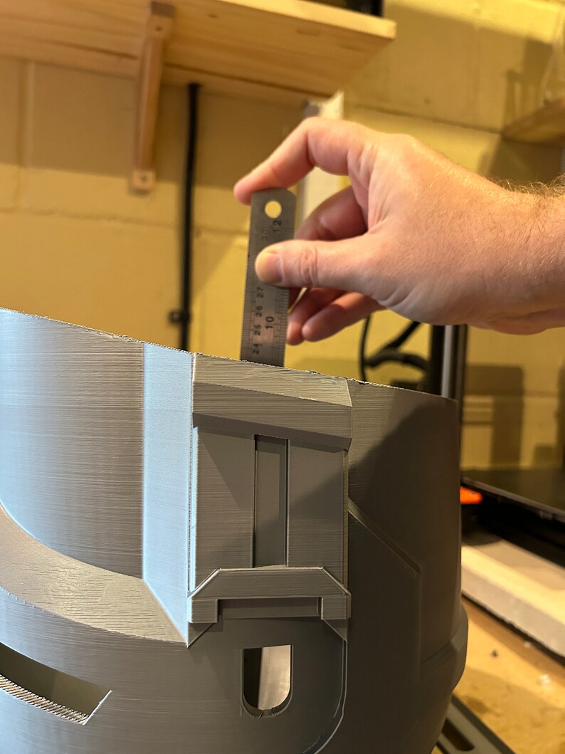 May include: A gray 3D printed helmet with a detailed design. A ruler is being used to measure the helmet, which is approximately 7 inches tall.