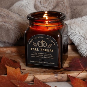 May include: A square amber glass candle jar with a black label featuring a pumpkin illustration and the text "CORDUROY LANE FALL BAKERY warm pumpkin cookies, sugared donuts, spiced coffee cakes, and sweet buttercream hand-poured soy | 8oz | 45 hours".