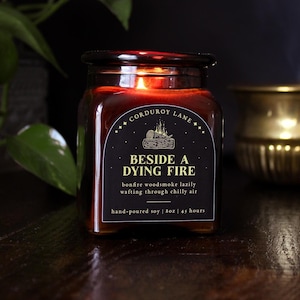 Peut inclure: Une bougie en pot de verre carré avec une étiquette noire portant le texte Corduroy Lane, Beside a Dying Fire, bonfire woodsmoke lazily wafting through chilly air, hand-poured soy, 227g, 45 hours. La bougie émet une lueur chaude de la mèche.
