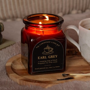 Peut inclure: Une bougie en verre ambré de forme carrée avec une étiquette noire portant le texte Corduroy Lane Earl Grey, a steaming mug of black bergamot tea with cream, hand-poured soy, 227g, 45 hours. La bougie est allumée et posée sur un plateau en bois.