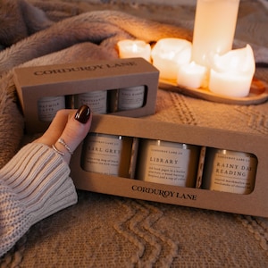 May include: Two cardboard boxes of scented candles from Corduroy Lane. One box contains three candles with labels that read "Earl Grey", "Library", and "Rainy Day Reading". The other box contains three candles with labels that read "Maymila Latte", "Croissant Honey", and "Fall Cabin".