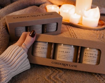 May include: Two rectangular cardboard boxes labeled CORDUROY LANE, each containing three glass jar candles. The front box features labels reading EARL GREY, LIBRARY, and RAINY DAY READING. The back box shows OATMILK LATTE, CROISSANTS & HONEY, and FALL BAKING.