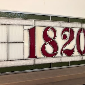 May include: A stained glass window with the number 1820 in burgundy red lettering on a white background. The window is framed in silver and has a green border.