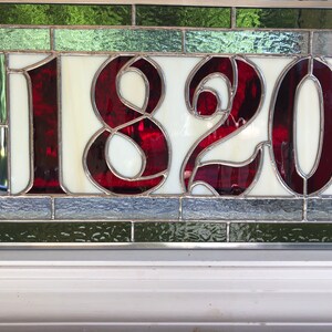 May include: A stained glass house number with the number 1820 in red on a white background. The glass is surrounded by a silver frame and green and gray glass.