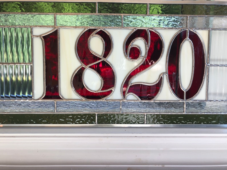 May include: A stained glass house number with the number 1820 in red on a white background. The glass is surrounded by a silver frame and green and gray glass.