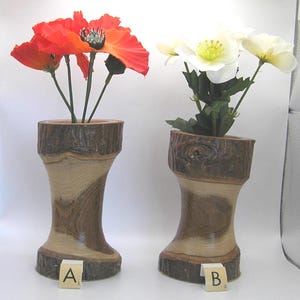 May include: Two wooden vases, each holding flowers. One vase has bright orange poppies, the other has white flowers. The vases are made of light brown wood with a natural bark edge. Each vase has a lettered tile at the base.