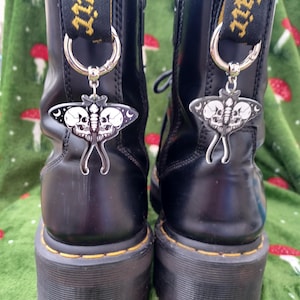 May include: Black platform boots with silver moth charms. The boots feature yellow stitching and black pull tabs with yellow text. The moth charms have a skull design and crescent moon details.