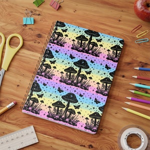 Könnte beinhalten: Ein spiralgebundenes Notizbuch mit einem bunten Regenbogenhintergrund und einem schwarzen Silhouettenmuster aus Pilzen und Sternen.