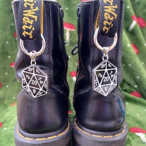 May include: Two black platform boots with silver D20 dice charms. The boots feature yellow and black pull tabs with "Dr. Martens" text. The dice charms are black and white.