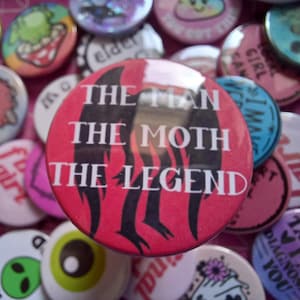 May include: A red button with the text "THE MAN THE MOTH THE LEGEND" in white. The button has a black and red flame design. Other buttons with various designs and text are in the background.