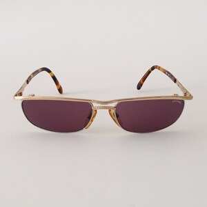 Puede incluir: Gafas de sol de metal dorado con montura de carey marr&oacute;n y lentes moradas. Las gafas tienen la inscripci&oacute;n 'COUNTY' en la patilla derecha.