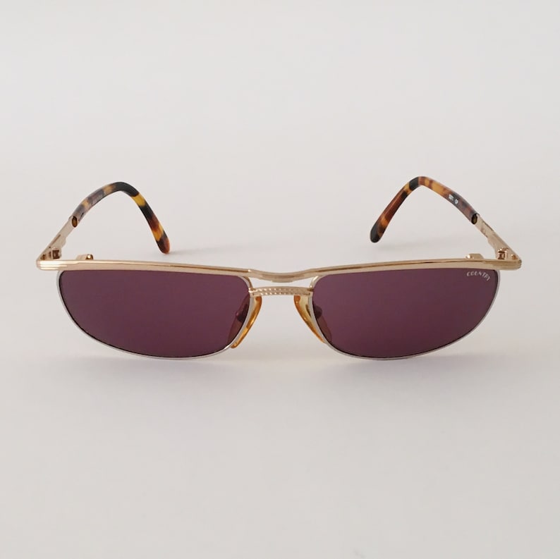 Puede incluir: Gafas de sol de metal dorado con montura de carey marr&oacute;n y lentes moradas. Las gafas tienen la inscripci&oacute;n 'COUNTY' en la patilla derecha.