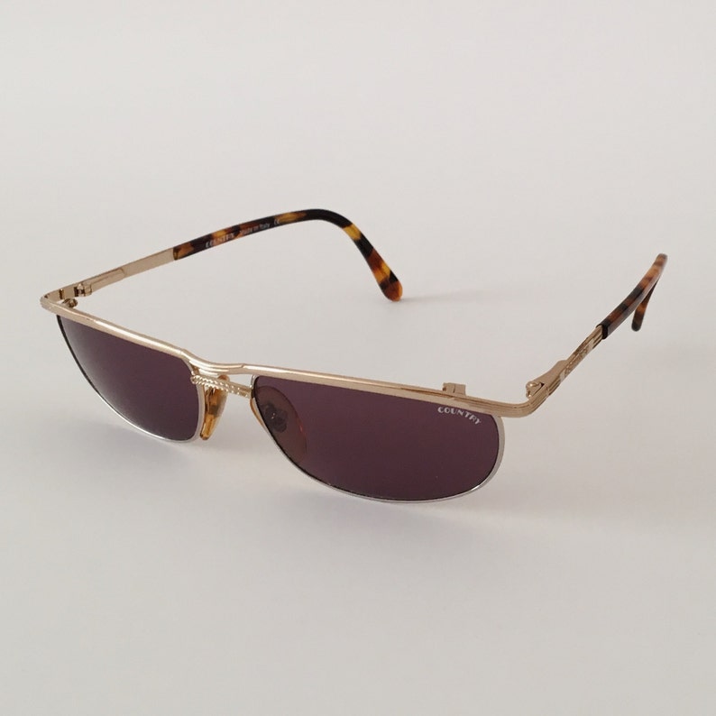 Puede incluir: Gafas de sol de metal dorado con patillas de carey marr&oacute;n y lentes grises oscuras. Las gafas tienen la palabra "COUNTER" impresa en la lente derecha.