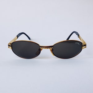 May include: Gold-rimmed oval sunglasses with brown frames and dark lenses. The sunglasses have the word "STING" written on the right lens.