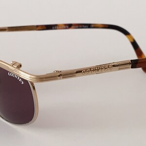 Puede incluir: Gafas de sol aviador de metal dorado con patillas de carey marr&oacute;n. Las gafas tienen lentes marrones oscuros y el nombre de la marca "COUNTRY" est&aacute; grabado en la montura.