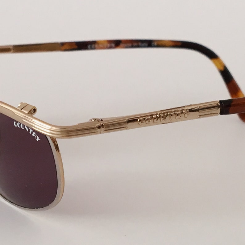 Puede incluir: Gafas de sol aviador de metal dorado con patillas de carey marr&oacute;n. Las gafas tienen lentes marrones oscuros y el nombre de la marca "COUNTRY" est&aacute; grabado en la montura.