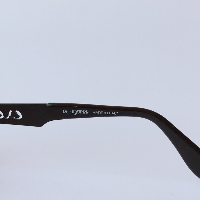 Peut inclure: Gros plan sur la branche en plastique marron d'une paire de lunettes. La branche est marqu&eacute;e "CE - EXESS - MADE IN ITALY".