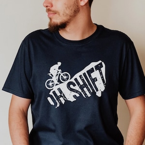 Könnte beinhalten: Ein marineblaues T-Shirt mit einer weißen Grafik einer Person, die mit dem Fahrrad über eine Klippe fährt, mit dem Text "OH SHIFT" in einer verwitterten Schrift.
