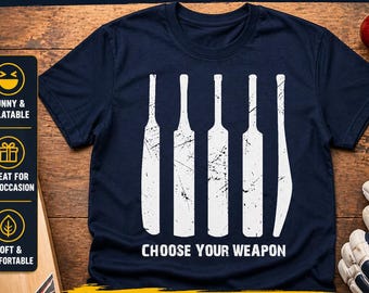 May include: Navy blue t-shirt with a white graphic of cricket bats and the text "CHOOSE YOUR WEAPON". The shirt also has the text "PERFECT GIFT FOR CRICKET LOVERS!" and "GAME DAY FAVORITE!". A cricket ball, helmet, and gloves are also visible.