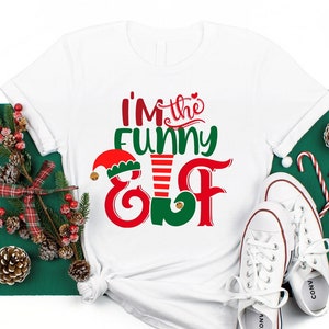 May include: White t-shirt with red and green text that reads "I'm the Funny Elf". The shirt is laying on a green surface with a Christmas tree branch, pine cones, and candy canes.