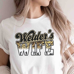 Peut inclure: T-shirt blanc avec l'inscription "Welder's WIFE" en noir et or avec un motif léopard. Le mot "WIFE" présente des silhouettes de soudeurs. Le t-shirt est porté avec un jean bleu et un cardigan crème.