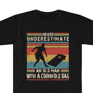 Op de afbeelding: Zwart T-shirt met een vintage-stijl grafische afbeelding met de tekst "Never underestimate an old man with a cornhole bag". De afbeelding toont een silhouet van een persoon die een beanbag gooit naar een cornhole-bord.