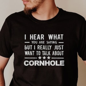 Puede incluir: Camiseta negra con texto blanco. El texto dice: "Escucho lo que dices, pero solo quiero hablar de cornhole". La camiseta tiene cuello redondo y mangas cortas. El texto está en negrita y sin serifa.