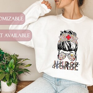 May include: White sweatshirt featuring a graphic of a woman with a messy bun wearing a nurse's hat and glasses. The text "Nurse Anesthetist" is printed below the graphic.