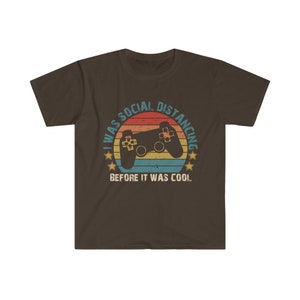 May include: A brown t-shirt with a retro-style graphic of a video game controller and the text "I was social distancing before it was cool."