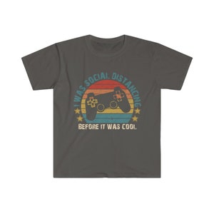 May include: A dark gray t-shirt with a retro-style graphic of a video game controller and the text "I was social distancing before it was cool."