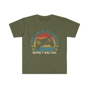 May include: A green t-shirt with a retro-style graphic of a video game controller and a sunset. The text reads "I was social distancing before it was cool."