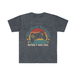 May include: A dark heather grey t-shirt with a retro-style graphic of a video game controller and the text "I was social distancing before it was cool."