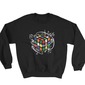 Puede incluir: Sudadera negra con un dise&ntilde;o de cubo de Rubik colorido, rodeado de flechas blancas y ecuaciones matem&aacute;ticas. El cubo tiene cuadrados rojos, naranjas, amarillos, verdes, azules y blancos. El dise&ntilde;o est&aacute; centrado en el pecho.