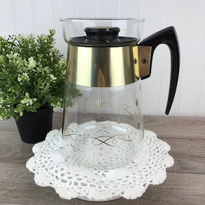May include: A clear glass coffee pot with a gold band and a black handle. The pot has a gold swirl design and sits on a white crocheted doily.