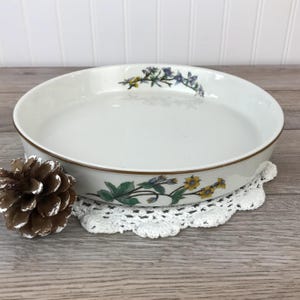 May include: A white ceramic baking dish with a floral design featuring yellow, blue, and green flowers. The dish has a brown rim and is sitting on a white crocheted doily.