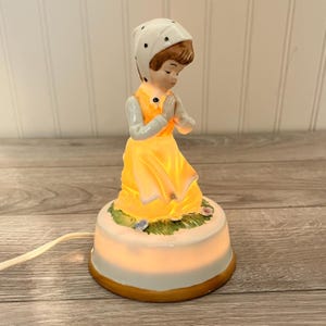 Peut inclure: Veilleuse en céramique représentant une fillette en robe jaune et coiffe blanche, les mains jointes en prière. La base ronde présente un motif floral et émet une lumière chaude. La lampe est reliée à un cordon électrique blanc. Hauteur environ 15 cm.