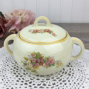 May include: A vintage cream-coloured sugar bowl with a lid. The bowl is decorated with a floral pattern in shades of pink, yellow, and green. The lid has a gold trim and a floral design.